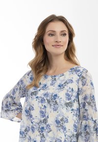 Blomsterbluse i let stof med blå, lilla og gule rosemønstre. Trekvart lange klokkeærmer og rund halsudskæring.