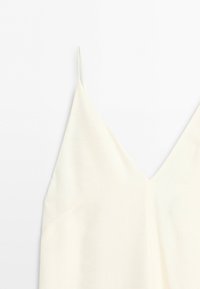 Close-up of a cream-colored sleeveless top with thin shoulder straps and a V-shaped neckline on a white background.