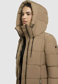 Pufferjack in tan met een hoge capuchon, voorzien van een rits aan de voorkant, trekkoordaccenten en een gequiltde textuur. Zwarte accenten zichtbaar.