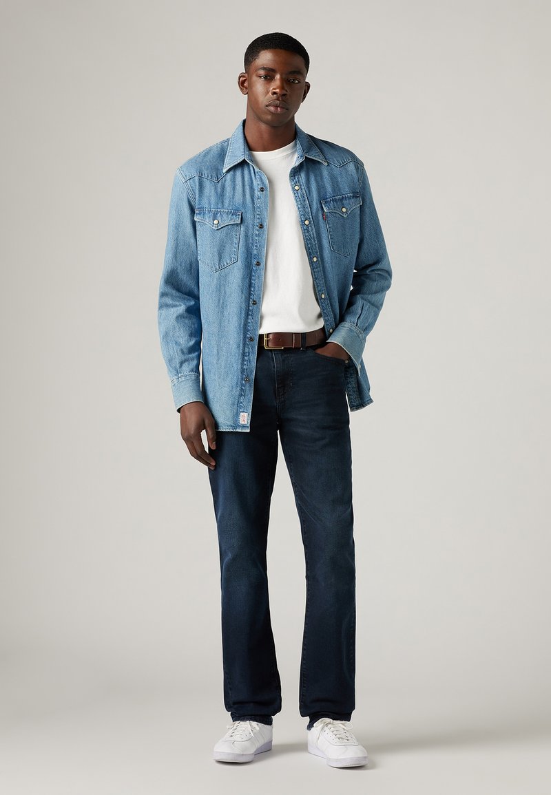 Camisa de mezclilla azul claro sobre una camiseta blanca, combinada con jeans de mezclilla oscura y zapatillas blancas. Cuenta con bolsillos abotonados y un corte relajado.