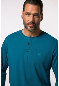 Tyrkysová dlouhá rukávová košile s kulatým výstřihem, tříknoflíkovou légou a malým logem tištěným na levé hrudi. Měkká textilie.