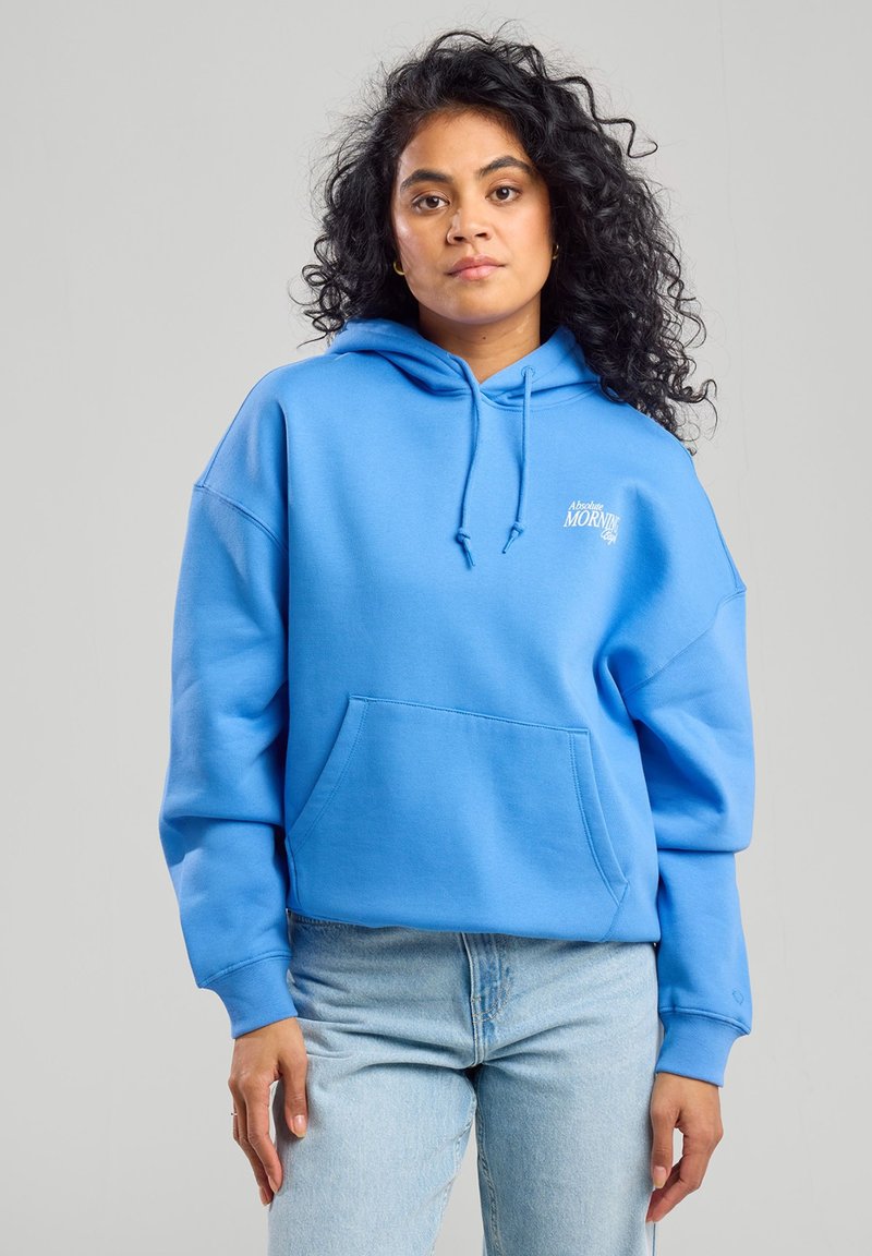 Blue hoodie with a kangaroo pocket, drawstring hood, and white text logo. Soft fabric, relaxed fit, and ribbed cuffs. Paired with light denim jeans.