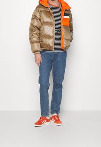 Homme portant une veste matelassée marron brillante avec une doublure orange, un t-shirt rayé noir et blanc, un jean bleu, et des baskets Adidas orange avec des rayures blanches.