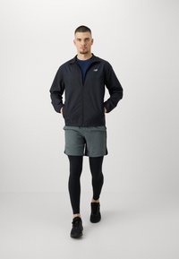 Sort zip-up jakke over en blå skjorte, parret med grå shorts og sorte leggings. Afsluttet med sorte sportssko.