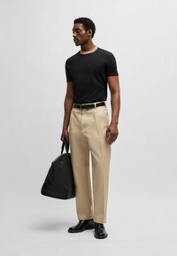 Maglietta nera a maniche corte con colletto beige, pantaloni beige a pieghe, cintura nera e scarpe di pelle nere; tiene in mano una borsa nera.