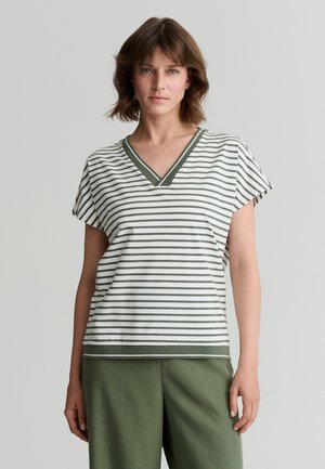 Femme aux cheveux courts bruns porte un chemisier blanc rayé vert à col en V et un pantalon vert olive, debout devant un fond gris clair.