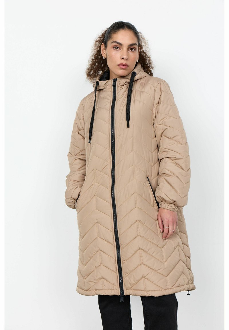 Femme portant une longue veste matelassée beige avec capuche et une fermeture éclair noire, debout devant un fond blanc uni.