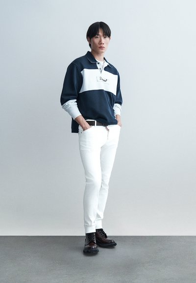 Jeune homme debout les mains dans les poches, portant une chemise manches longues bleu marine et blanche, un pantalon blanc et des bottes marron foncé sur un sol gris.