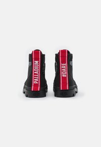 Stivaletti neri alla caviglia con strisce laterali rosse con il testo "#DARE" e "PALLADIUM". Suola spessa e tessuto testurizzato con dettagli del logo.