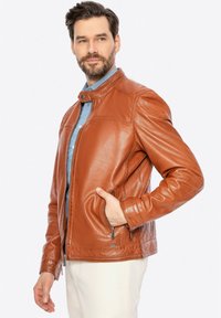 Giacca in pelle marrone con colletto alto, frontale con zip e due tasche laterali. Texture liscia, design aderente e dettagli di cucitura sottili.