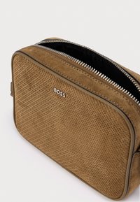 Καφέ τσάντα από σουέτ με υφή, στρογγυλεμένες άκρες, ασημένιο φερμουάρ και μικρό λογότυπο στο μπροστινό μέρος. Το εσωτερικό είναι μαύρο.