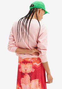 Sudadera rosa clara ajustada en la cintura, combinada con una falda de patrón abstracto multicolor. Una gorra verde adorna la cabeza.