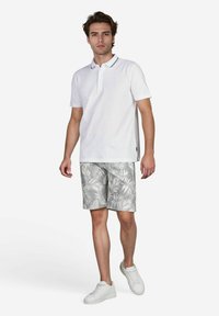 Weißes Poloshirt mit Kragen, kombiniert mit grauen Shorts im Blattmuster. Modell trägt weiße Sneakers und steht vor einem weißen Hintergrund.