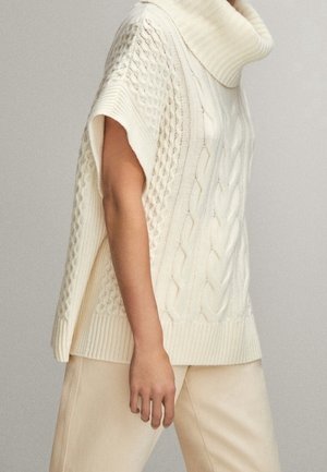 Roomkleurige mouwloze kabelgebreide coltrui gedragen met bijpassende lichtbeige broek tegen een effen achtergrond.