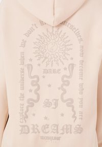 Sudadera de color rosa claro con un gran gráfico texturizado en la parte posterior que presenta un sol, un símbolo de yin-yang, serpientes y texto estilizado.