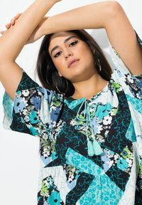 Kukallinen tunika, jossa on sinisen, vihreän ja mustan kuvion sekoitus. Rennonmallinen, jossa on nauha kaula-aukossa ja tupsukoristeita. Lyhyet hihat.