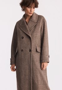 Brązowy wełniany płaszcz dwurzędowy z szerokim kołnierzem, dużymi guzikami, bocznymi kieszeniami i lekko teksturowanym materiałem. Długi, luźny krój.