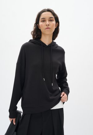 Felpa nera con cappuccio e cordini, realizzata in materiale morbido. Presenta una vestibilità comoda e spacchi laterali, indossata sopra una maglietta bianca.