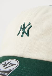 Gorra de béisbol con un cuerpo de algodón crema y visera verde. Presenta un logotipo bordado en verde. Incluye un parche circular con detalles de la marca.
