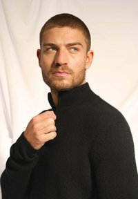 Czarny sweter wykonany na drutach z wysokim kołnierzem. Modelka ma krótkie włosy i podnosi kołnierz jedną ręką. Gładka faktura i dopasowany krój.