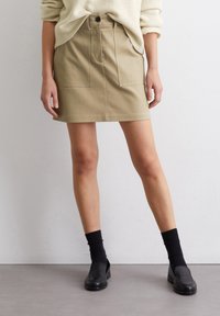 Mini jupe en tissu khaki avec poches avant, taille haute et détails de couture. Portée avec des chaussettes noires mi-mollet et des chaussures à enfiler.