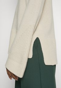 Krämmönstrad stickad tröja med sidsspringa och långa ribbade ärmar, bar över mörkgröna byxor på en persons arm.