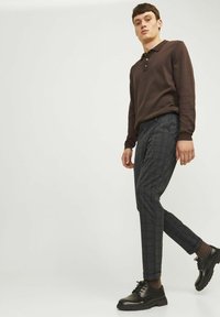 Dunkelbraunes Langarm-Polo-Shirt mit Knöpfen, kombiniert mit grauen karierten Hosen und schwarzen Schnürschuhen mit einer klobigen Sohle.