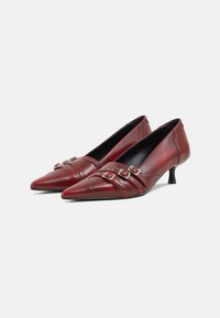 Zapatos de tacón de cuero rojo con punta afilada, con detalles de hebilla y costuras horizontales, y un pequeño tacón negro.