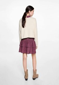 Beige verkürzter Pullover über einem geschichteten, rüschigen, burgunderfarbenen Rock mit kleinen Blumenmustern. Trägt braune Ankle Boots mit kleinen Absätzen.
