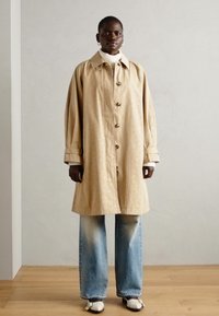 Bežs garais mētelis ar spīdīgu tekstūru, pogu priekšpusi un plašām piedurknēm, kurš valkāts virs baltā turtlenecka, saskaņots ar izbalējušām ziloņa biksēm un melnām kurpēm.