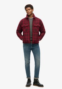 Chaqueta de cuadros roja y negra con dos bolsillos en el pecho, llevada sobre un suéter gris, combinada con jeans azules y botas negras con cordones.