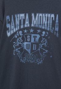 Tengerészeti kék pamut póló, melyen nagy, fakó grafikával szerepel a "SANTA MONICA" felirat, csillagok és egy pajzs dizájn a "1991" évszámmal.