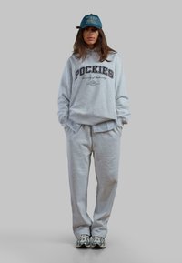 Szary bawełniany hoodie z napisem "POCKIES", zestawiony z dopasowanymi szarymi spodniami dresowymi. Na głowie niebieska czapka z logiem, noszona tyłem do przodu.