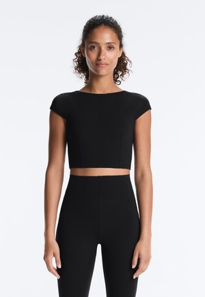 Kvinde med krøllet hår iført en tætsiddende sort crop top og sorte leggings med høj talje, stående mod en ensfarvet hvid baggrund.