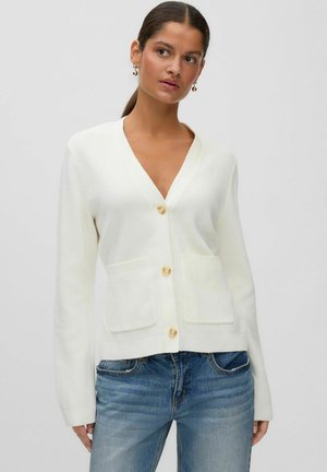 Femme aux cheveux foncés portant des boucles d'oreilles en or, un cardigan blanc boutonné avec des poches avant et un jean bleu, debout devant un fond uni.
