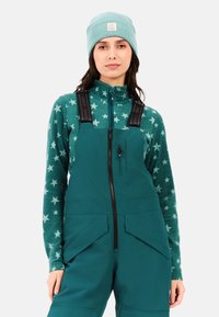 Teal ski jumpsuit met verstelbare zwarte banden, gecombineerd met een teal fleece top met witte sterpatronen, en een bijpassende gebreide beanie.