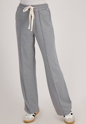 Vrouw in een grijze brede joggingbroek met een witte trekkoord taille en witte sneakers met zwarte strepen.