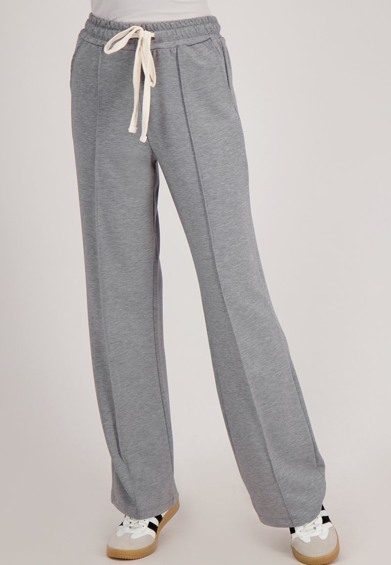Vrouw in een grijze brede joggingbroek met een witte trekkoord taille en witte sneakers met zwarte strepen.