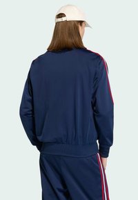 Námornícka tréningová bunda s červenými bočnými pruhmi, vyrobená z hladkej tkaniny. Obsahuje rebrovaný pás a manžety. Čiapka nosená naopak.