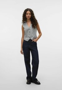 Grå ärmlös skjorta med knappstängning och V-ringning, kombinerad med mörkblå vida jeans. Modellen står med en hand i fickan.