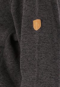 Grå texturerad sweatshirtärm med en beige läderlogotyp med skölddesign. Tyget verkar mjukt och något melerat.