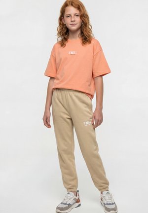 Roodharig kind draagt een oranje O'Neill T-shirt, beige O'Neill sweatpants en witte sneakers, staand tegen een eenvoudige lichte achtergrond.