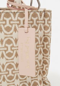 Beige en bruine handtasstof met een patroon, voorzien van een lichtroze leren label dat is geperst met gouden tekst, en een bijpassende strap.