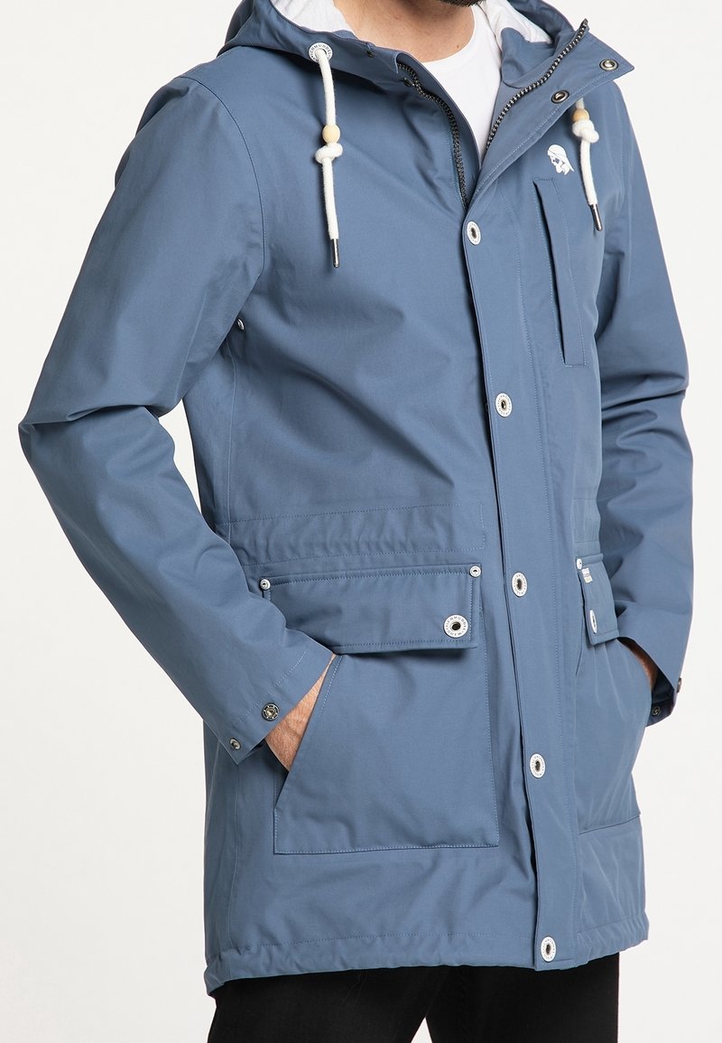 Blå vattentålig jacka med huva, knapp- och dragkedjeslutning framtill, två stora framfickor och dragsnoddar som accent. Slät textur.