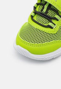 Zapatilla atlética de malla verde con cordones negros, tejido texturizado, puntera reforzada y suela de goma blanca con patrones de agarre.