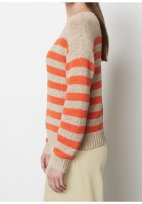 Kobieta w beżowym i pomarańczowym paskowanym swetrze dzianinowym oraz jasnobeżowych spodniach, stojąca bokiem na jednolitym białym tle.