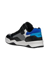 Zapatilla deportiva en negro y gris con una correa de velcro, suela blanca y detalles azules en el talón y la lengüeta. Presenta tejidos texturizados y cuero suave.