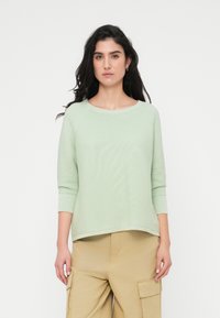 Ανοιχτό πράσινο, πλεκτό πουλόβερ με στρογγυλεμένο λαιμόκοψη και μανίκια τριών τετάρτων, σε συνδυασμό με χακί παντελόνι cargo, φορεμένο από μοντέλο.