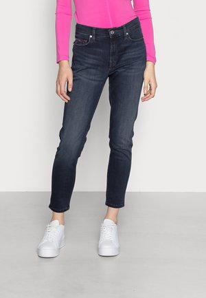 Person wearing dark blue slim-fit jeans, white sneakers, and a bright pink long-sleeve top, standing on a light gray floor.