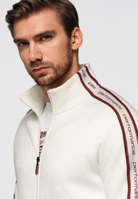 Jeune homme aux cheveux courts bruns et barbe, portant une veste blanche zippée avec le texte "performance" sur des bandes bordeaux le long des épaules.